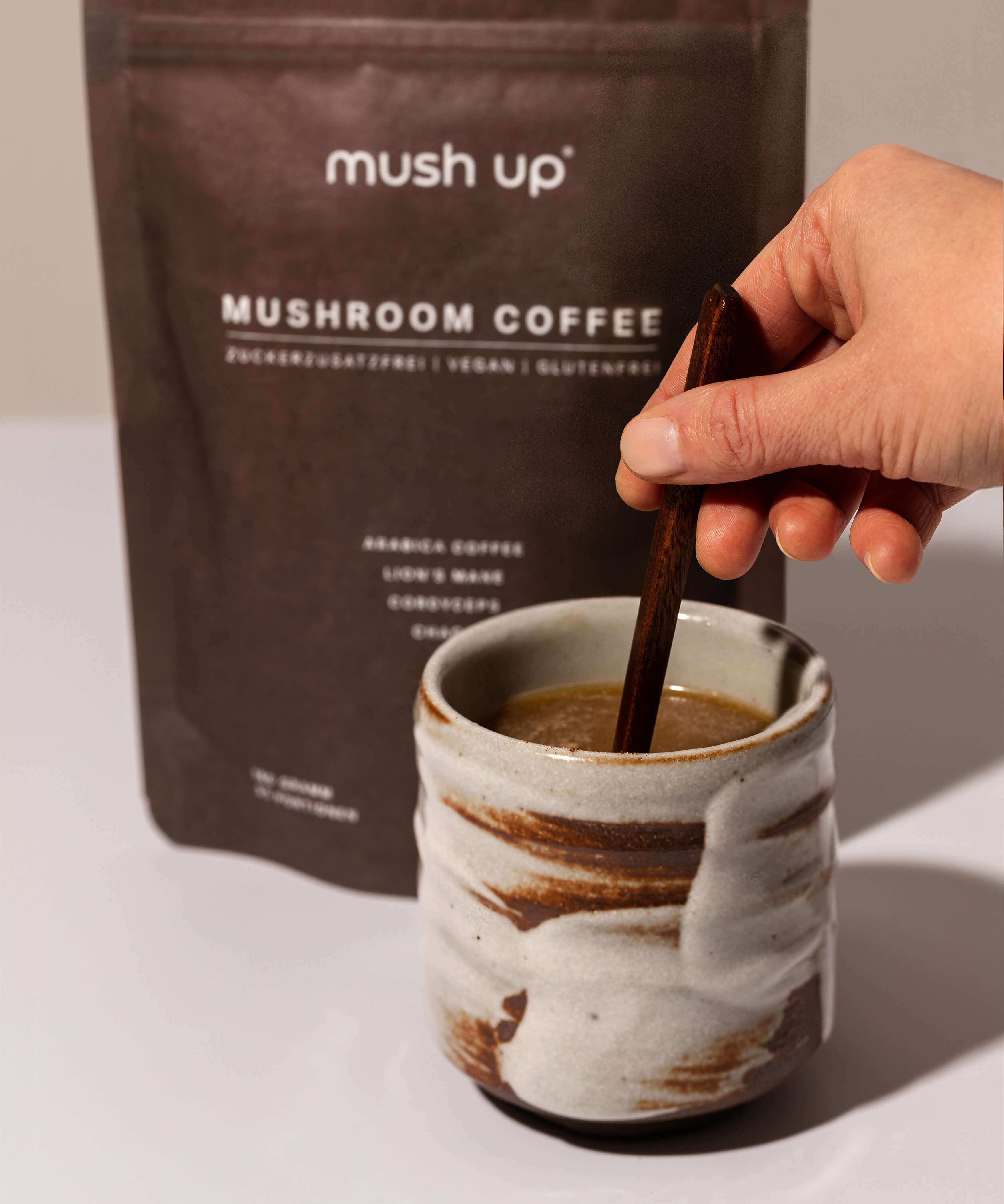 Zubereitung von mush up Mushroom Coffee – eine Hand verrührt das Pulver mit einem Holzlöffel in einer handgefertigten Keramiktasse, im Hintergrund die Packung mit Lion’s Mane, Chaga und Cordyceps Vitalpilzen.