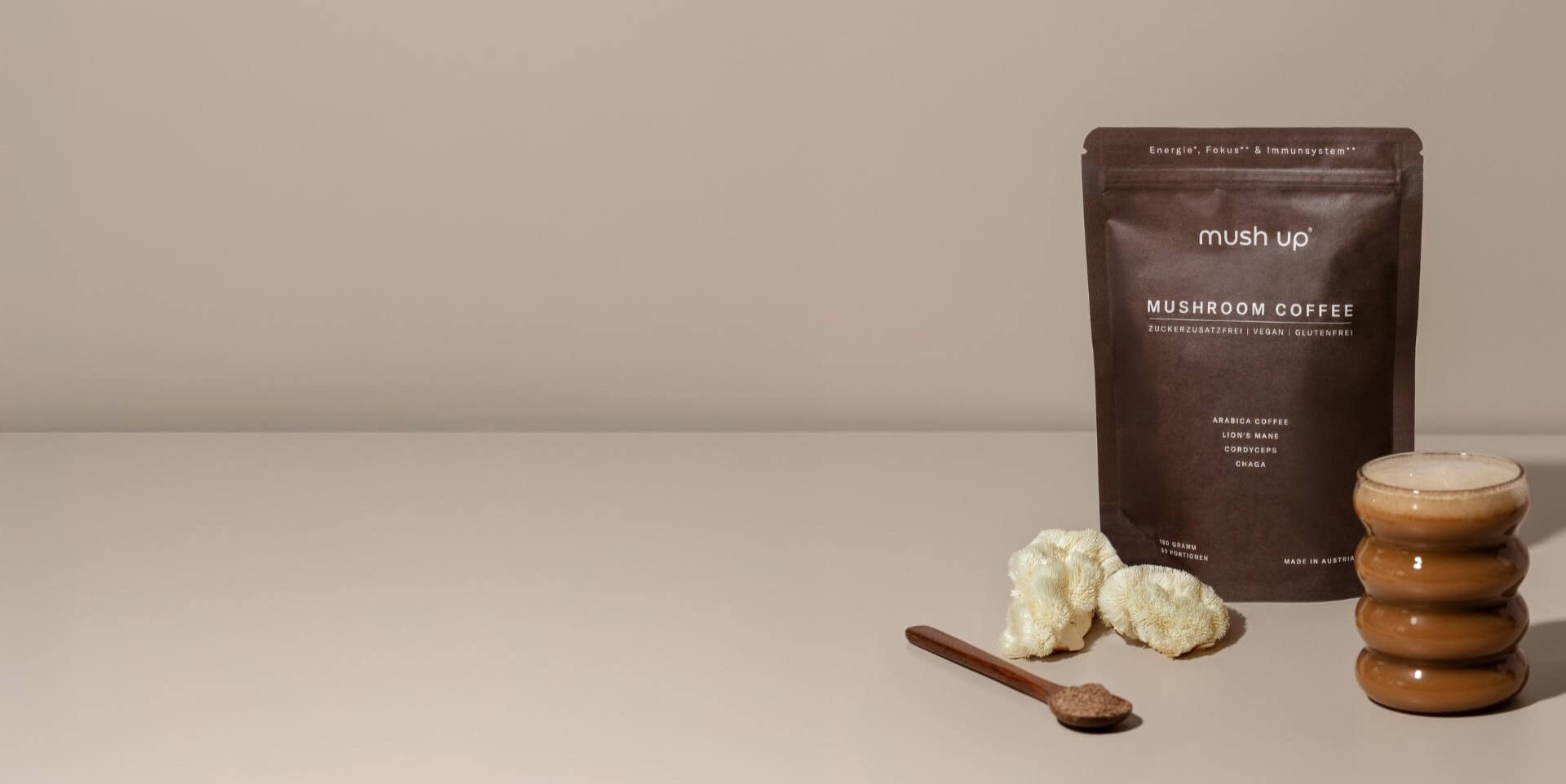 Edles Produktfoto von Mushroom Coffee von mush up – braune Verpackung mit Vitalpilz-Kaffeemischung, daneben der Heilpilz Lion's Mane, eine moderne Kaffeetasse mit cremigem Schaum und ein Akazien Holz-Löffel.