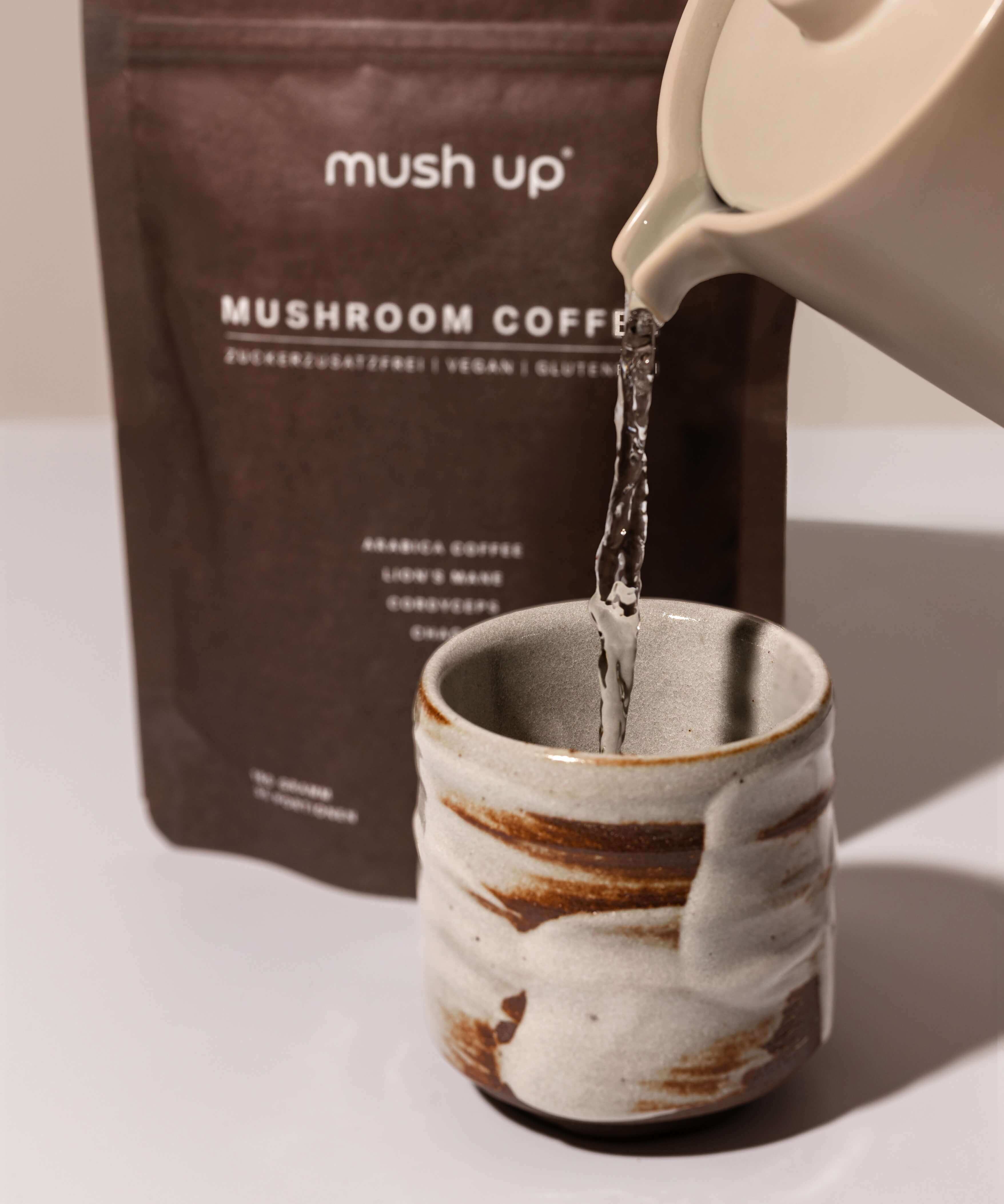 Zubereitung von mush up Mushroom Coffee – heißes Wasser wird aus einer Karaffe in eine handgefertigte Keramiktasse gegossen, im Hintergrund die Packung mit Lion’s Mane, Chaga und Cordyceps Vitalpilzen.