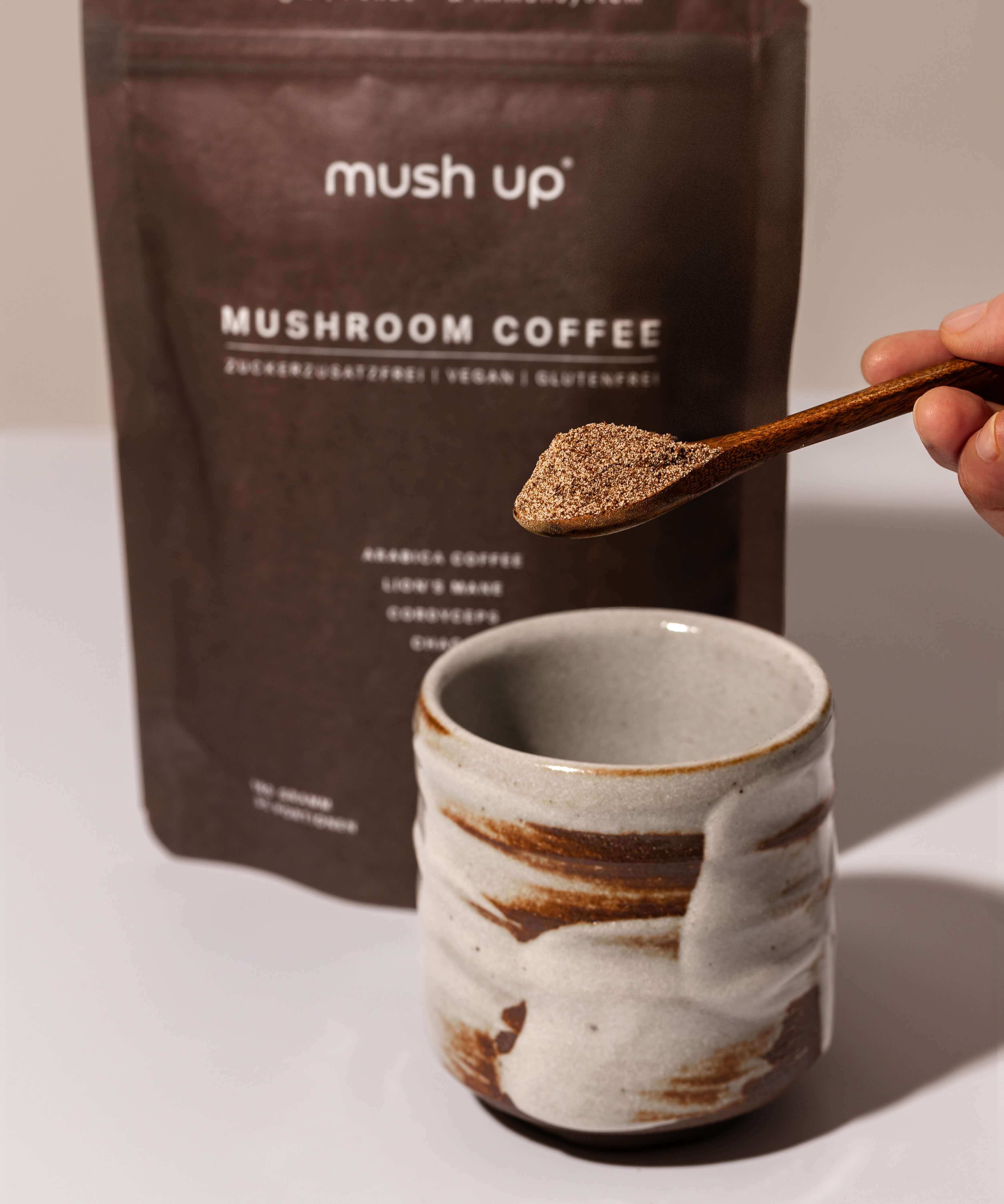 Zubereitung von mush up Mushroom Coffee – ein Holzlöffel mit Pulver wird in eine handgefertigte Keramiktasse gegeben, im Hintergrund die Packung mit Lion’s Mane, Chaga und Cordyceps Heilpilzen.